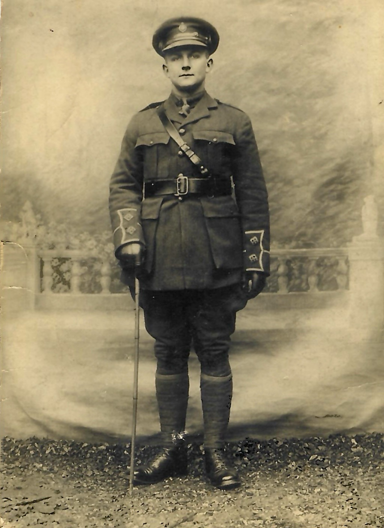 Soldier in full uniform, brandishing a cane by his right side, stands at attention in front of the painted canvas background of a balcony.