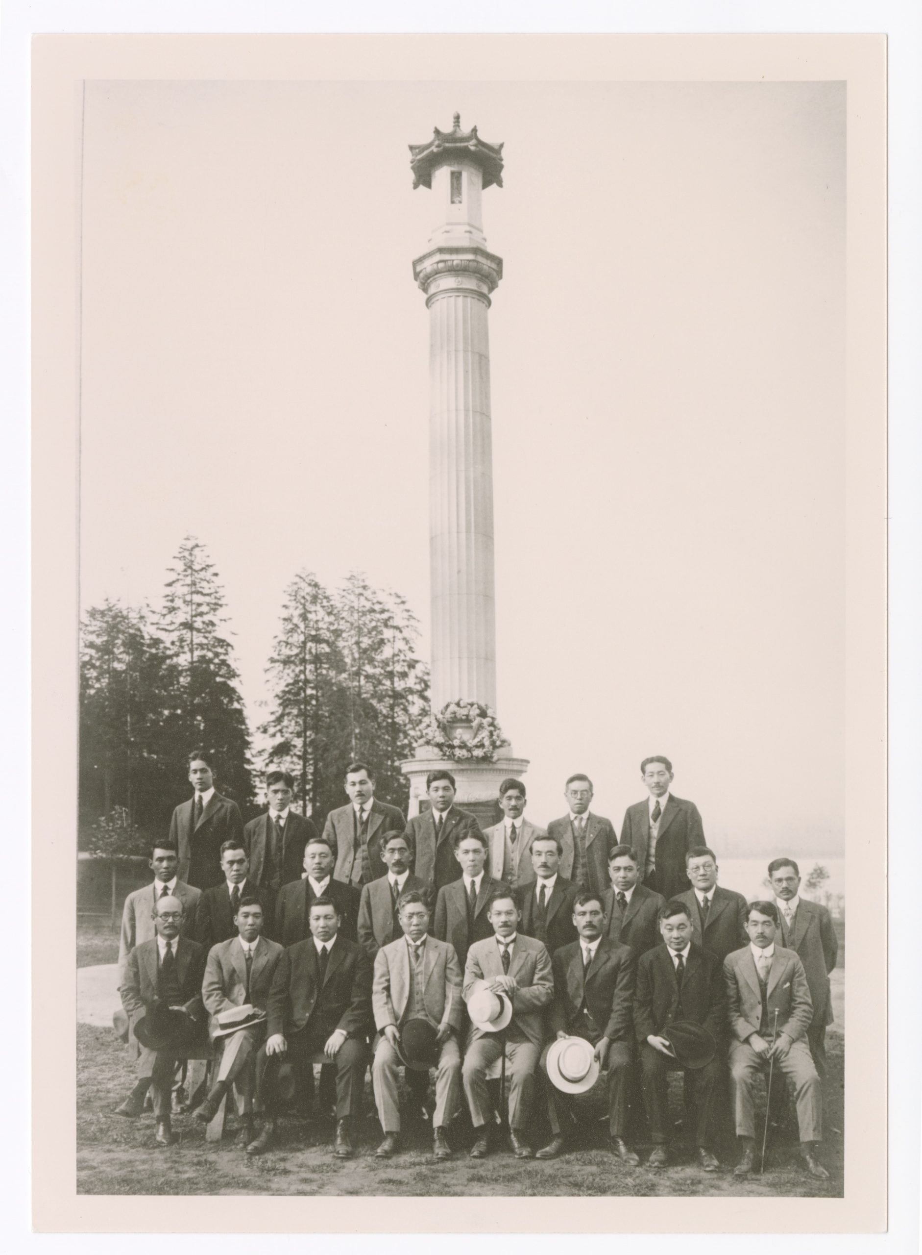 21 men assembled in 3 rows, dressed in suits stand in front of a monumental column that sports a wreath at its base.