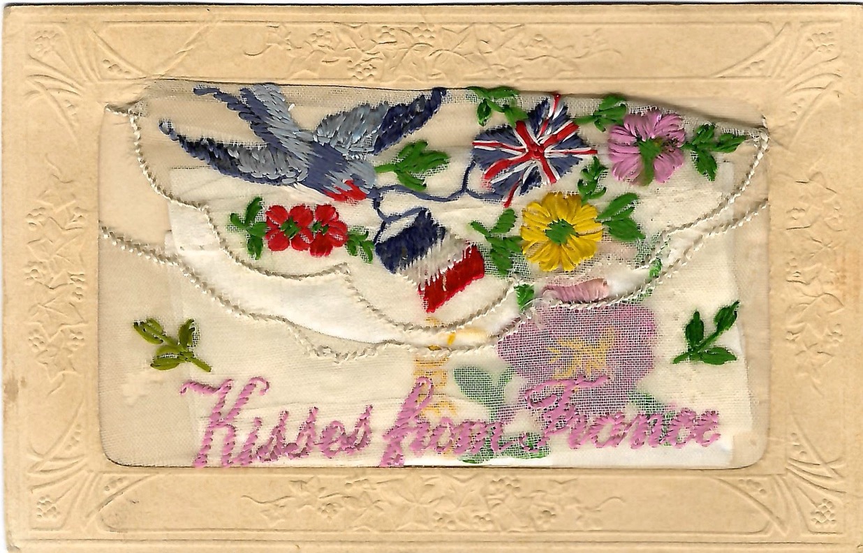 Mail card with fabric stitching of bird carrying vines abloom with red and yellow flowers and the flags of the United Kingdom and France. Written below is “Kisses from France.”