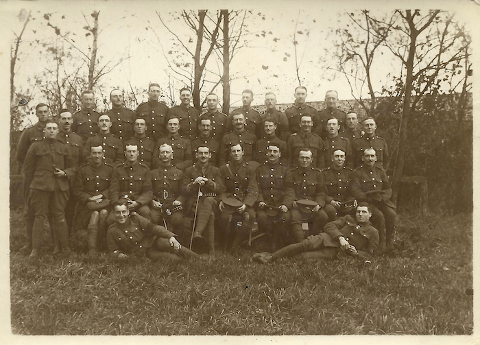 A group of soldiers pose on grass, while two lay on either flank. Behind them are hedges and a grouping of leafless trees.