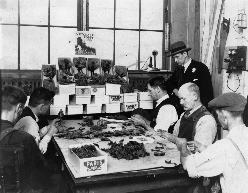 5 men sit around a table assembling poppies, while completed piles lay in the middle and foreground. Boxes lay stacked in the background.