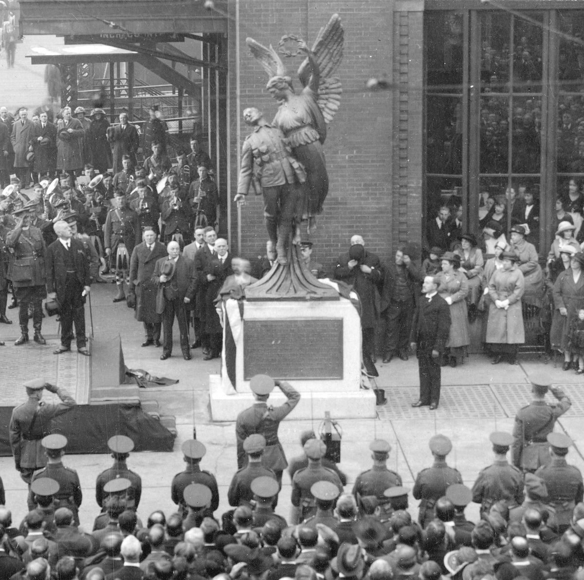 Crowds of civilians and servicemen gather around a monument in front of a brick building, some are seen saluting.