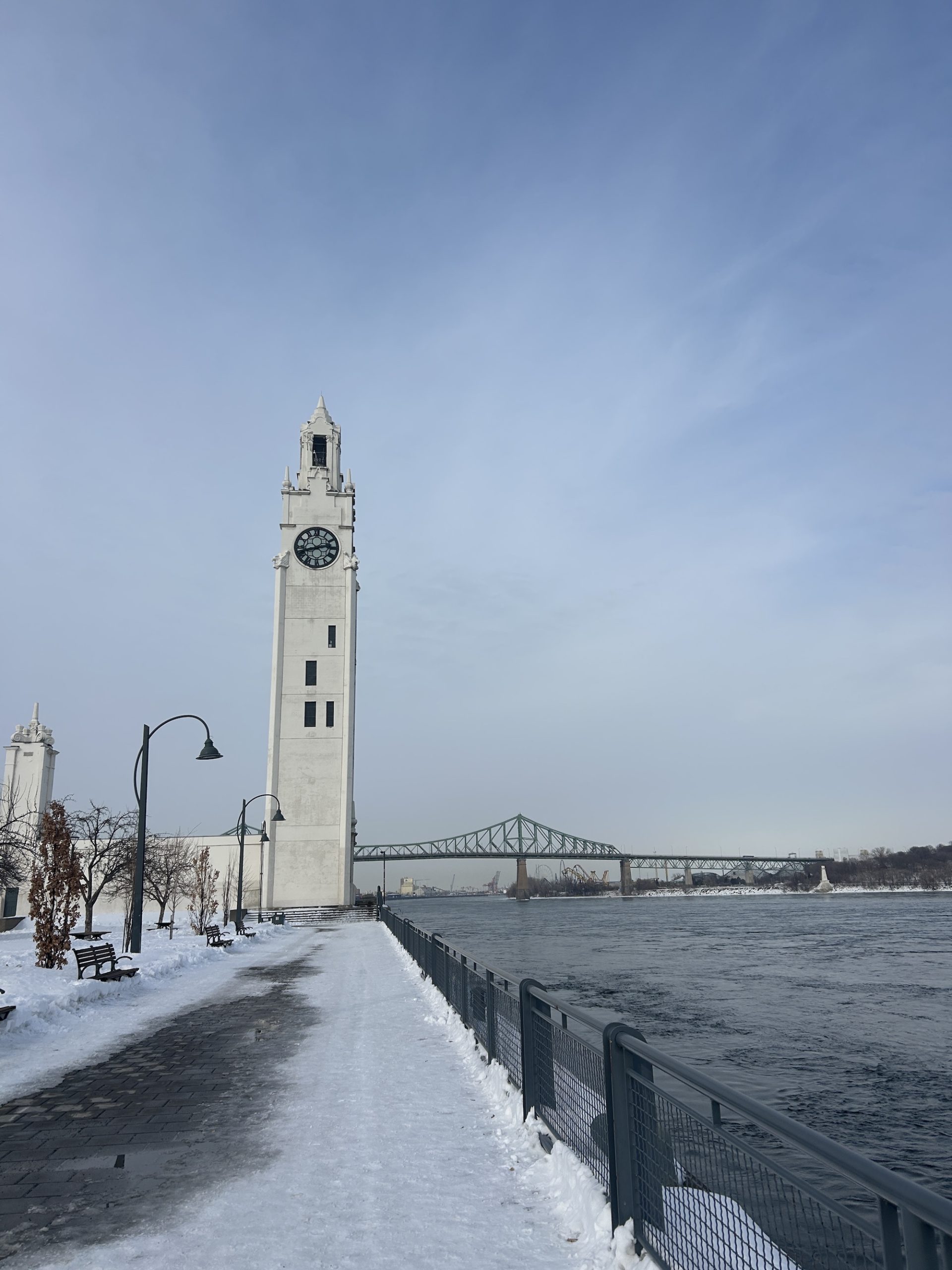 A snowy pathway, flanked on the right by water, leads to a lone ivory white clock tower. A steel bridge lays in the background.