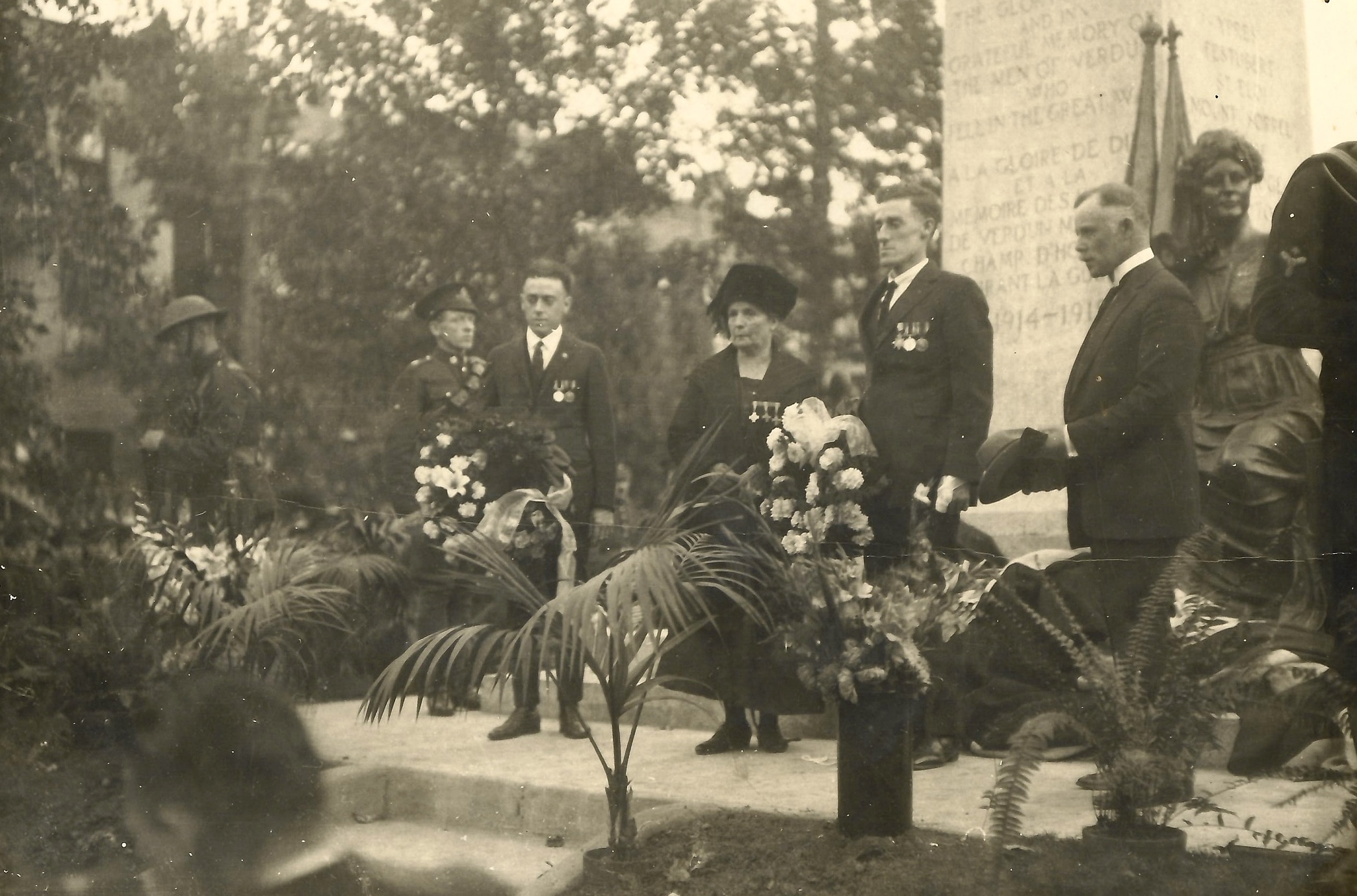 A group of two men and one woman sporting several medals stand in front of an engraved pillar and statuette, sporting solemn expressions gaze of into the distance with various potted plants and flowers placed at their feet. Soldiers take watch in the background and foreground.