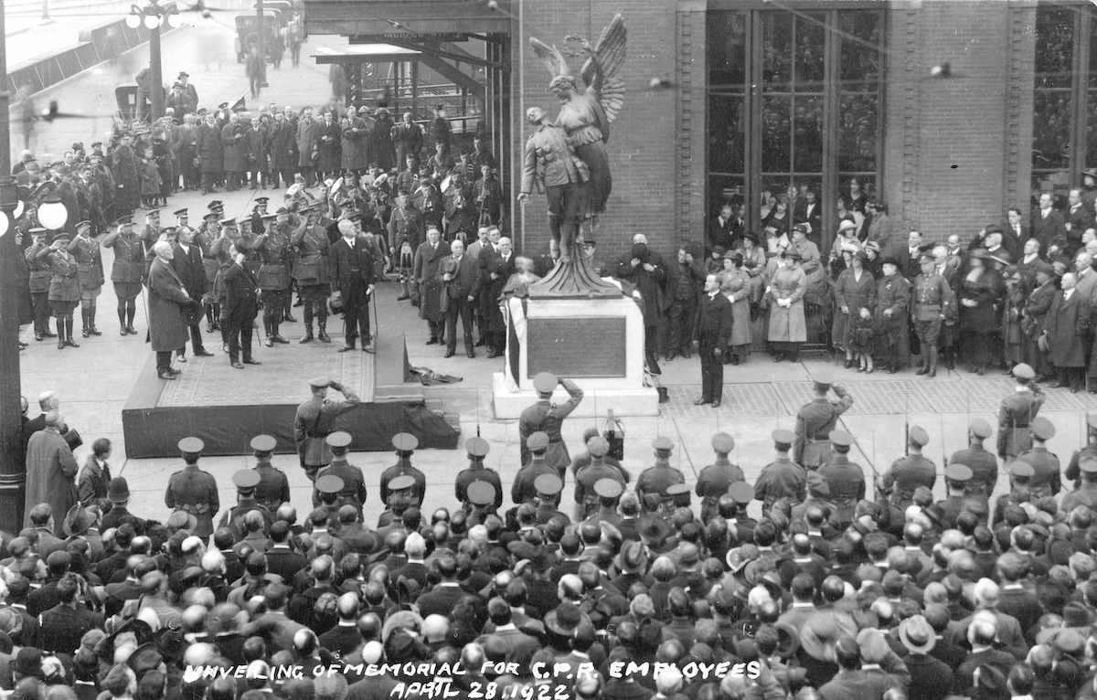 Crowds of civilians and servicemen gather around a monument in front of a brick building, some are seen saluting.